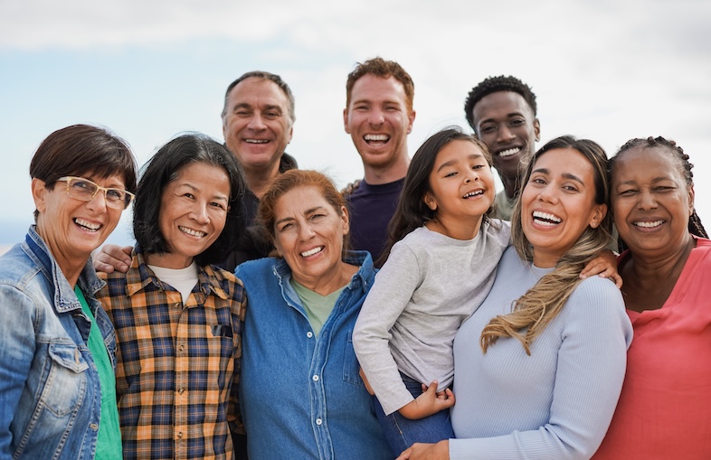 A diverse group of people, spanning different ages and ethnicities, standing together outdoors. They are smiling warmly and embracing each other, conveying a sense of unity and community. Among them is a young girl being held by a woman, with everyone appearing happy and connected.